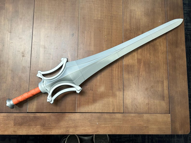 May include: A silver sword with a long, pointed blade and a detailed hilt. The handle is wrapped in orange material. The sword is lying on a wooden surface.