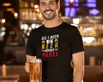 Może przedstawiać: Czarny t-shirt z napisem "ALL I NEED IS BEER BEFORE PADEL" w kolorze białym, żółtym i czerwonym. Osoba trzyma dużą szklankę piwa. Tło to bar z jedzeniem i napojami na stole.