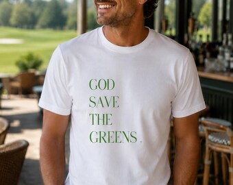 Może przedstawiać: Biała koszulka z zielonym napisem "GOD SAVE THE GREENS". Koszulka jest noszona przez osobę w okularach przeciwsłonecznych, stojącą przed polem golfowym i barem.