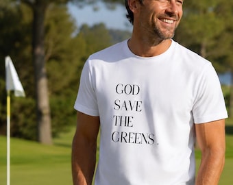 Może przedstawiać: Biała koszulka z czarnym napisem "GOD SAVE THE GREENS". Osoba uśmiecha się na polu golfowym, w tle drzewa i flaga golfowa. Koszulka jest głównym elementem.