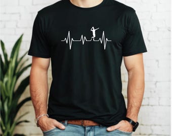 Może przedstawiać: Czarny t-shirt z białym nadrukiem przedstawiającym siatkarza wkomponowanego w linię bicia serca. Projekt sugeruje pasję do siatkówki. Ciemny materiał nadaje się do noszenia na co dzień.