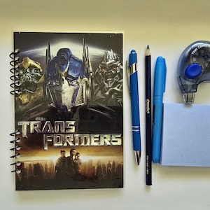 May include: A spiral-bound notebook featuring a Transformers movie graphic with Optimus Prime, a blue retractable pen, a black pencil, a blue marker, a blue correction tape dispenser, and a square light blue sticky note pad.