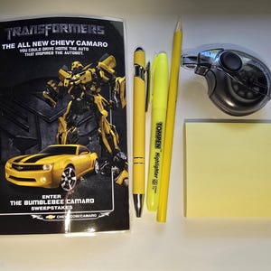 May include: A spiral-bound notebook featuring a Transformers Bumblebee graphic, a yellow ballpoint pen, a yellow Tompen highlighter, a yellow pencil, a black correction tape dispenser, and a square yellow sticky note pad.
