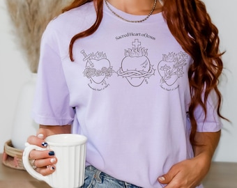 May include: A light purple t-shirt featuring the text "Sacred Heart of Jesus" above three heart illustrations. Each heart has a different design: a sword and flowers, a crown of thorns, and flowers. The shirt is a soft, comfortable material.