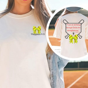 Puede incluir: Camiseta color crema con un dise&ntilde;o de lazo de softbol y el texto "Todo lo puedo en Cristo que me fortalece" en la espalda. La parte delantera tiene un dise&ntilde;o de lazo m&aacute;s peque&ntilde;o y el texto "Filipenses 4:13."