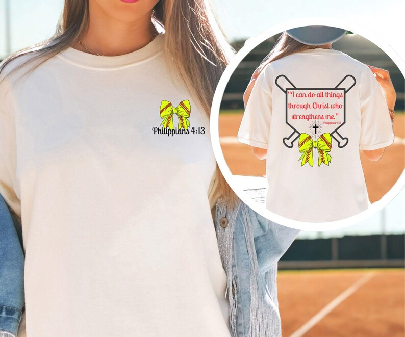 Puede incluir: Camiseta color crema con un dise&ntilde;o de lazo de softbol y el texto "Todo lo puedo en Cristo que me fortalece" en la espalda. La parte delantera tiene un dise&ntilde;o de lazo m&aacute;s peque&ntilde;o y el texto "Filipenses 4:13."
