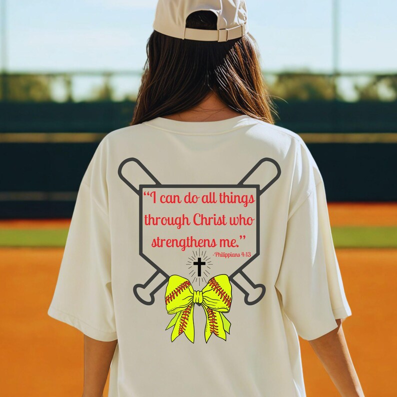 Puede incluir: Camiseta color crema con un dise&ntilde;o de softbol en la espalda. El dise&ntilde;o incluye bates de b&eacute;isbol cruzados, una pelota de softbol y un lazo amarillo. El texto dice: "Todo lo puedo en Cristo que me fortalece." Filipenses 4:13.