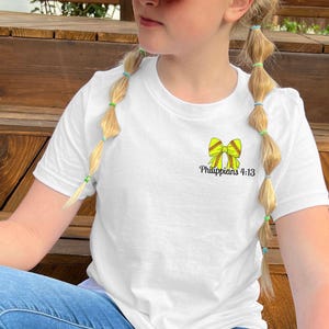 Puede incluir: Camiseta blanca con un lazo de softbol amarillo y el texto "Filipenses 4:13". La camiseta se combina con vaqueros azules. La persona lleva una gorra de b&eacute;isbol roja y blanca.