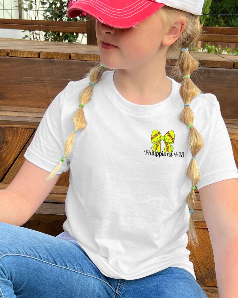 Puede incluir: Camiseta blanca con un lazo de softbol amarillo y el texto "Filipenses 4:13". La camiseta se combina con vaqueros azules. La persona lleva una gorra de b&eacute;isbol roja y blanca.