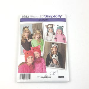 May include: Simplicity pattern 1953 featuring animal-themed hats. The pattern includes designs for a fox, owl, monkey, frog, and panda hat. The cover shows photos of people wearing the hats. The pattern size is A/S-L/L.