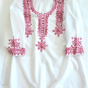 May include: A white long-sleeved tunic featuring a scoop neckline and intricate embroidery in shades of pink and magenta. The geometric needlework patterns are concentrated around the neckline, down the center front, and on the sleeve cuffs.
