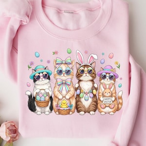 May include: A pale pink sweatshirt with a graphic of four cats celebrating Easter. The cats are wearing sunglasses, hats, and bunny ears. They are holding Easter baskets and are surrounded by decorative eggs and flowers.