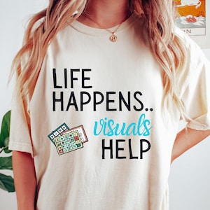 Peut inclure: T-shirt crème avec l'inscription "LIFE HAPPENS.. visuals HELP" en noir et turquoise. Un petit graphique de calendrier est imprimé sur le devant. Vêtement de style décontracté.