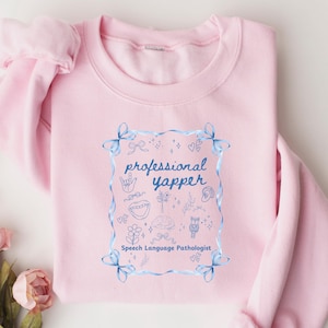 Puede incluir: Sudadera rosa claro con la frase "professional yapper" en un marco azul. El diseño incluye ilustraciones y las palabras "Speech Language Pathologist". La sudadera tiene cuello redondo y mangas largas.