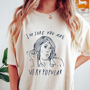 以下が含まれることがあります： 「I'M SURE YOU ARE VERY POPULAR」の文字と女性の黒い線画が描かれたクリーム色のTシャツ。カジュアルなスタイルで、普段使いに適しています。