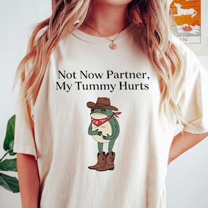 May include: Cream-colored t-shirt with the text "Not Now Partner, My Tummy Hurts." The shirt features a cartoon frog wearing a cowboy hat, red bandana, and boots. The frog is holding its stomach.