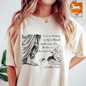 Puede incluir: Camiseta color crema con una ilustración en blanco y negro de una escena bíblica y texto. El texto dice: "si te aferras a un hilo, asegúrate de que sea el borde de su vestidura" y "Mateo 9:20-27".
