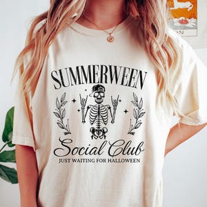 May include: Cream-colored t-shirt with the text "SUMMERWEEN Social Club" and a skeleton graphic. The design includes a skeleton wearing a cap, surrounded by leaves and stars. The phrase "JUST WAITING FOR HALLOWEEN" is below.