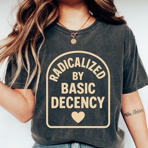Pode incluir: T-shirt cinza escuro com o texto "RADICALIZED BY BASIC DECENCY" em um design arqueado bege, com um coração abaixo. A camisa é combinada com jeans azuis e tênis brancos. O tema é roupa casual.