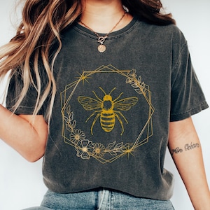 May include: Dark gray t-shirt featuring a gold bee design within a geometric frame with floral accents. The bee is detailed with stripes and wings. The shirt is paired with blue jeans and white shoes, creating a casual, everyday look.