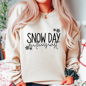 May include: A beige sweatshirt with the text "SNOW DAY enthusiast" in black, decorated with snowflake graphics. The sweatshirt is paired with plaid pants. The overall theme is winter and cozy apparel.