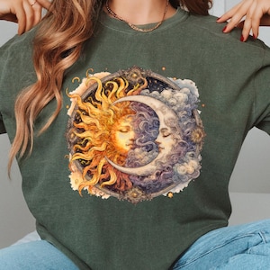May include: Dark green t-shirt with a detailed illustration of the sun and moon, each with a serene face, set within an ornate circular frame. The sun is depicted in warm yellow and orange hues, while the moon is in cool blues and purples.