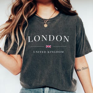 Puede incluir: Camiseta gris oscuro con la palabra "LONDON" en blanco, una bandera británica y "UNITED KINGDOM" debajo. La camiseta se combina con vaqueros azules y zapatillas blancas. Tema: ropa de viaje y urbana.