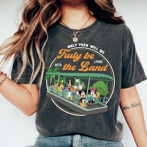 Könnte beinhalten: Dunkelgraues T-Shirt mit einer Grafik eines Boots mit Disney-Figuren und dem Text "Only Then Will We Truly be Living with the Land." Das Shirt wird mit blauen Jeans und weißen Turnschuhen getragen.