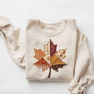 May include: A cream-coloured crewneck sweatshirt featuring a large, central maple leaf appliqué. The leaf is constructed from various fabric patches in shades of brown, orange, and tan, showcasing diverse patterns including plaid, floral, and solid textures.