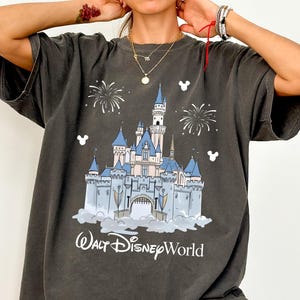 May include: Dark grey t-shirt with a graphic of Cinderella's castle, blue turrets, fireworks, and the text "Walt Disney World." The shirt has short sleeves and a relaxed fit, suitable for casual wear.