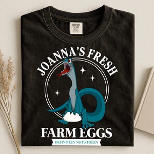 Puede incluir: Camiseta negra con un dibujo animado de dinosaurio. El dise&ntilde;o presenta un dinosaurio azul con una lengua roja, el texto "JOANNA'S FRESH FARM EGGS" en un dise&ntilde;o circular y la frase "DEFINITELY NOT STOLEN" debajo.