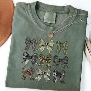 May include: An olive green t-shirt featuring nine bow designs in various camouflage patterns. A gold necklace with a pendant rests on the shirt. The shirt is folded, with a brown hat and dried plant stems in the background.