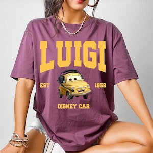 May include: A purple t-shirt featuring the word "LUIGI" in yellow block letters above a cartoon car graphic. The graphic includes the text "EST 1959 DISNEY CAR". The shirt is short-sleeved.
