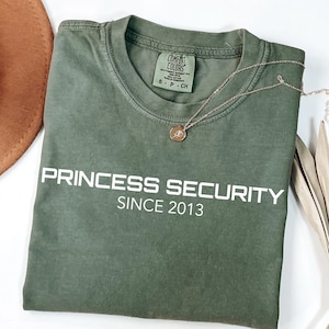 Może przedstawiać: Oliwkowo-zielona koszulka z białym napisem "PRINCESS SECURITY SINCE 2013". Złoty naszyjnik z wisiorkiem jest zarzucony na koszulkę. Brązowy kapelusz i suszona roślina znajdują się w tle.