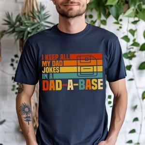 May include: Navy blue t-shirt with the text "I KEEP ALL MY DAD JOKES IN A DAD-A-BASE" in a retro color scheme. The design includes a graphic of a floppy disk. A casual tee for a dad.