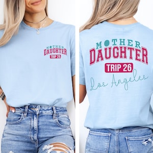 May include: Light blue t-shirt with the text "MOTHER DAUGHTER TRIP 26" in pink and white on the front and back. The back of the shirt also has "Los Angeles" in a teal script font. The model is wearing blue jeans.