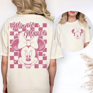 Puede incluir: Camiseta color crema con un gr&aacute;fico de Minnie Mouse rosa. La parte posterior de la camiseta muestra una cara grande de Minnie Mouse con un lazo, sobre un fondo a cuadros rosa y blanco. La parte delantera tiene un gr&aacute;fico de Minnie Mouse m&aacute;s peque&ntilde;o.