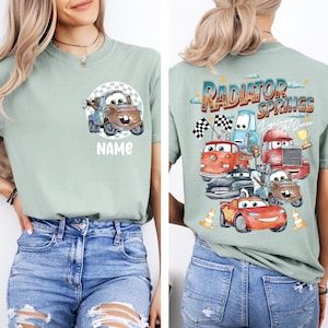 May include: Sage green t-shirt featuring a cartoon image of Mater from the movie Cars, with the word "NAME" below. The back of the shirt displays a graphic of various Cars characters with the text "Radiator Springs Lightning McQueen".