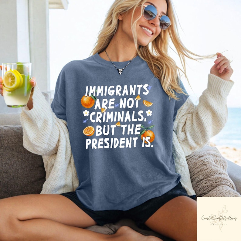 May include: A blue t-shirt with the text "IMMIGRANTS ARE NOT CRIMINALS, BUT THE PRESIDENT IS." Oranges and flowers are printed around the text. The shirt is worn by a person wearing sunglasses and holding a drink.