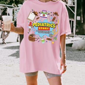 May include: A light pink t-shirt featuring a colorful graphic print with medical-themed illustrations, including a stethoscope, bandages, and a rocket. The text reads PEDIATRICS STORY TO INFINITY AND BEYOND FOR OUR PATIENTS in yellow and blue.