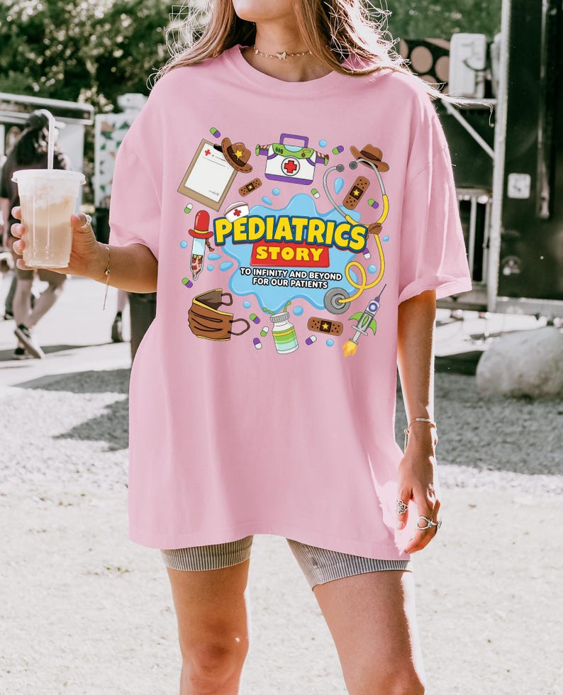 May include: A light pink t-shirt featuring a colorful graphic print with medical-themed illustrations, including a stethoscope, bandages, and a rocket. The text reads PEDIATRICS STORY TO INFINITY AND BEYOND FOR OUR PATIENTS in yellow and blue.