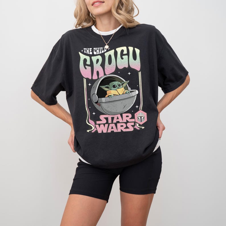 Puede incluir: Una camiseta negra de manga corta con un estampado gr&aacute;fico de Grogu en una c&aacute;psula flotante. El texto dice THE CHILD GROGU y STAR WARS en una fuente de estilo retro con colores rosa y verde claro. Tambi&eacute;n se ve un peque&ntilde;o icono de casco mandaloriano.