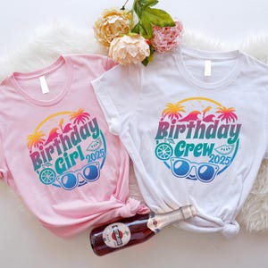 May include: Two short-sleeved t-shirts, one pink and one white, with a colorful graphic that reads "Birthday Girl 2025" and "Birthday Crew 2025". The design includes sunglasses, palm trees, and a slice of watermelon. A bottle of sparkling wine is in the image.