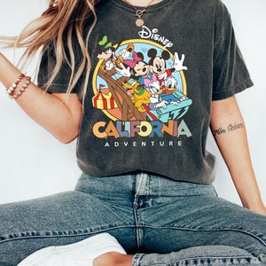 May include: Dark gray t-shirt featuring a colorful graphic of Disney characters on a ride, with the words "Disney California Adventure" in a circular design. The shirt is worn with blue jeans and white sneakers.