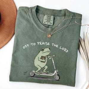 Può includere: T-shirt verde oliva con la scritta "OFF TO PRAISE THE LORD" e un ranocchio dei cartoni animati su uno scooter, con un libro in mano. Una collana d'oro è drappeggiata sulla maglietta.