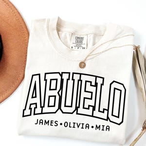 May include: Cream-colored t-shirt with the word "ABUELO" in bold, black, block letters. Below the word are the names JAMES, OLIVIA, and MIA. A gold necklace and a brown hat are also visible.