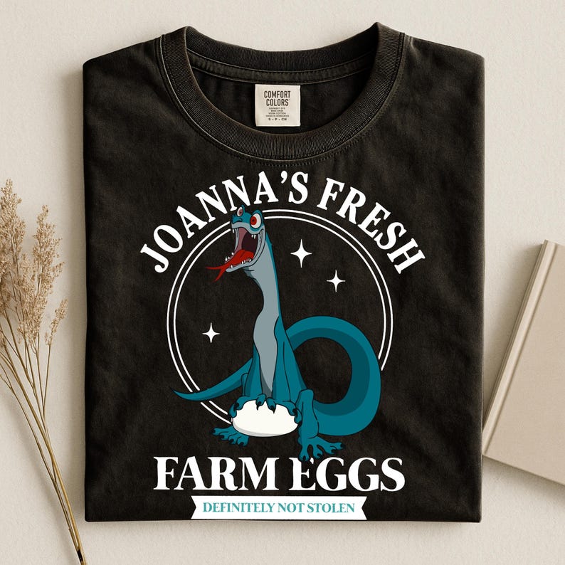 Puede incluir: Camiseta negra con un dibujo animado de dinosaurio. El dise&ntilde;o presenta un dinosaurio azul con una lengua roja, el texto "JOANNA'S FRESH FARM EGGS" en un dise&ntilde;o circular y la frase "DEFINITELY NOT STOLEN" debajo.