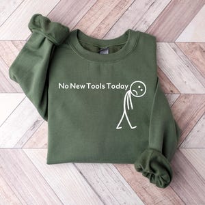 Puede incluir: Sudadera verde oliva con el texto "No New Tools Today" y un dibujo de un hombre de palitos. La sudadera está doblada sobre una superficie clara con vetas de madera.