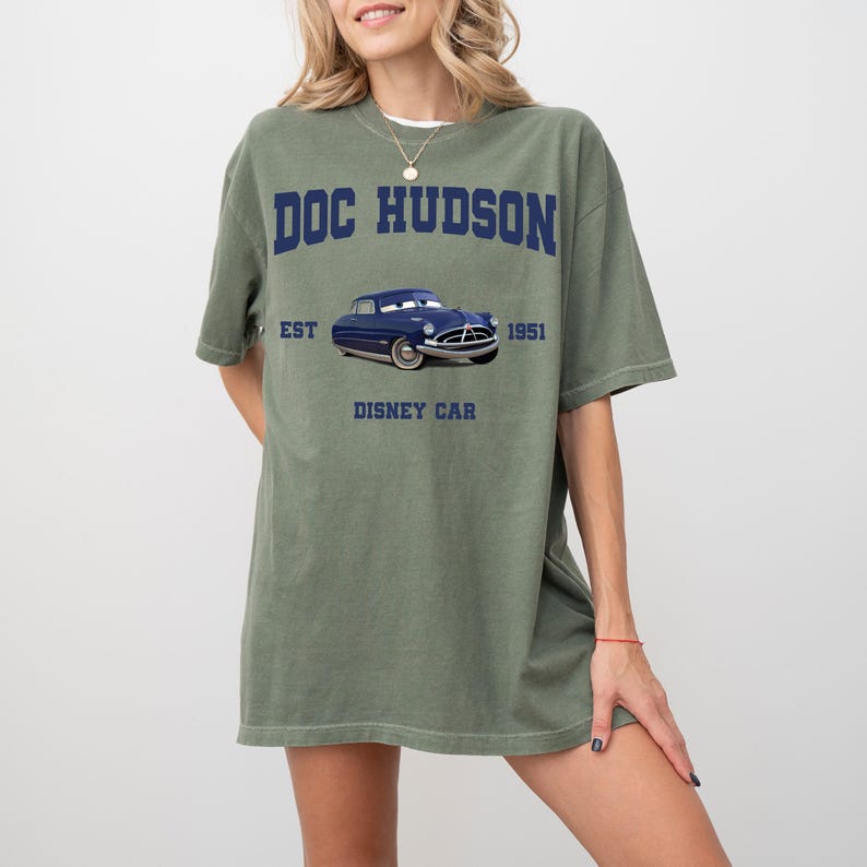 May include: Olive green t-shirt featuring the text "DOC HUDSON" above a blue cartoon car graphic. Below the car are the words "EST 1951 DISNEY CAR". The shirt has short sleeves and a relaxed fit.