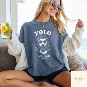 Peut inclure: Un t-shirt bleu avec le mot "YOLO" au-dessus d'un dessin blanc de J&eacute;sus portant des lunettes de soleil. Le texte "J/K, BRB - JESUS" est en dessous. Le t-shirt est port&eacute;.